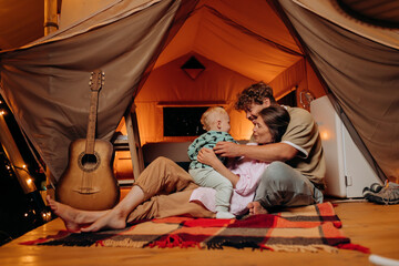 Fototapeta na wymiar Happy family with lovely baby playing and spend time together in glamping on summer evening. Luxury camping tent for outdoor recreation and recreation. Lifestyle concept