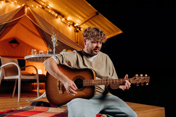 Obraz premium Handsome young smiling man playing guitar sitting in cozy glamping tent in summer evening bonfire. Luxury camping tent for outdoor holiday and vacation. Lifestyle concept