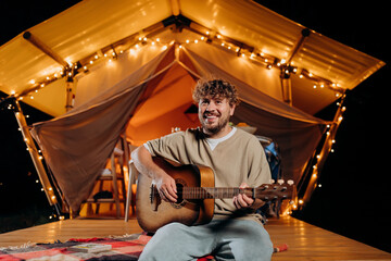 Obraz na płótnie Canvas Handsome young smiling man playing guitar sitting in cozy glamping tent in summer evening bonfire. Luxury camping tent for outdoor holiday and vacation. Lifestyle concept
