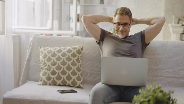 Older man working online with laptop computer at home sitting on couch in living room. Home office, browsing internet. Portrait of happy, mature age, middle age, mid adult man in 50s, smiling.