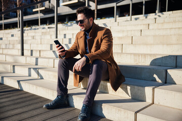 Happy stylish man wearing coat sitting on stairs in the city using phone