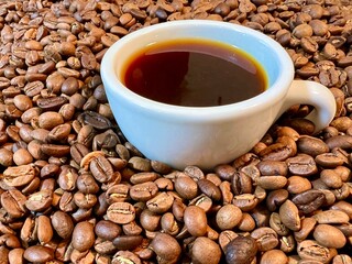 A white cup stands buried or plunged into a large pile of coffee beans It is filled with dark coffee