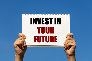 Invest in your future text on notebook paper held by 2 hands with isolated blue sky background. This message can be used as business concept about investing in your future.