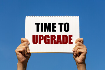 Time to upgrade text on notebook paper held by 2 hands with isolated blue sky background. This message can be used as business concept to inform audience that it is time for upgrading.