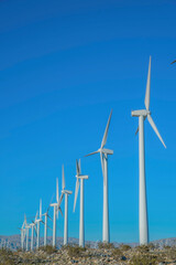 White wind turbines on a desert land in California. Low angle view of windmills with tubular steel tower against the mountain range and blue sky background.