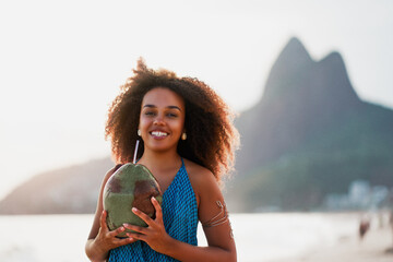 headshot portrait young black brazilian woman posing and smiling holding coconut water on the shore of the beach in Ipanema Brazil - obrazy, fototapety, plakaty