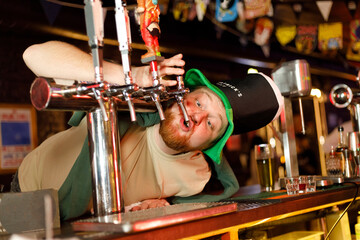 red-haired guy in a leprechaun hat for st. patrick's day drinks beer from a tap on a bar counter