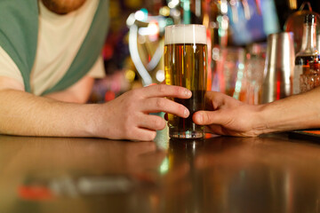 Mug of beer for client. Bartender puts light lager on wooden brown bar counter