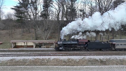 A Side Drone View of a Steam Locomotive Approaching Traveling Thru Fields and Meadows, Blowing White Smoke on a Winter Day