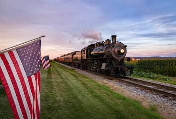 A View of a Classic Steam Passenger Train Approaching, With American Flags Attached to a Fence on a Sunny Summer Day