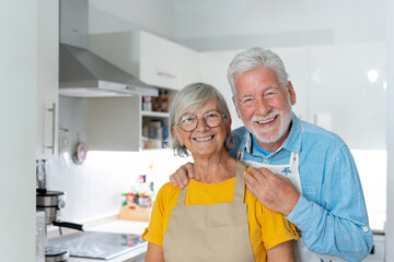 Head shot portrait smiling mature married couple looking at camera, standing in kitchen cooking at...