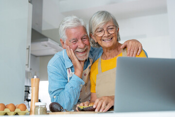 Happy joyful older husband and wife sharing cooking looking laptop, making organic fresh salad together, following recipe, talking, laughing. Old couple having fun in kitchen, preparing dinner.