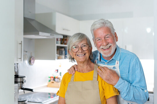 Head shot portrait smiling mature married couple looking at camera, standing in kitchen cooking at home, happy senior man wearing glasses hugging wife, grandparents posing for photo.