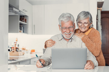 Old age and modern tech. Retired family couple sit at kitchen table use laptop watch photo video...
