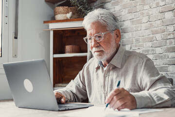 Fototapeta premium Aged remote worker. Concentrated senior male in glasses work on laptop from home office read email electronic document. Old age man employee freelancer sit at kitchen table by pc typing report online.