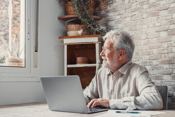 Smiling mature 70s man in glasses sit at table working on laptop look in distance enjoy happy...