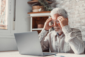 Tired mature 70s man working at laptop from home, taking off glasses, touching head and eyes. Old...