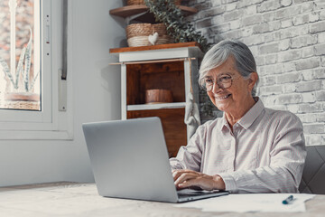 Aged remote worker. Concentrated senior female in glasses work on laptop from home office read email electronic document. Old age woman employee freelancer sit at kitchen table by pc typing report 