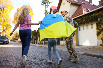 Soldier in military uniform with his family running and holding Ukrainian flag on city street, back...