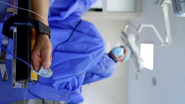 Male patient’s hand with attached sensor and tied to the handrail. Close up. Operating surgeon at backdrop in blur. Vertical view.