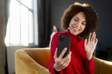 Portrait smiling African American woman holding mobile phone communication online, waving hand, having video call sitting on armchair at home. Influencer recording video using smartphone. Social media