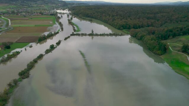 AERIAL: Rising muddy river overflowing its banks and flooding the countryside. Flooded landscape after abundance of rain in autumn season. Floodwater spreading over grassy riverside, fields and woods.