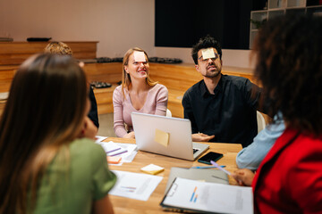 Diverse happy excited coworkers playing guessing game with sticky notes on forehead after work for entertainment. Cheerful multiracial group of colleagues having fun together with charades in office.