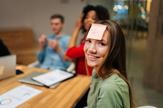 Close-up face of cheerful young woman playing guessing game with sticky notes on forehead after work for entertainment with multiethnic happy excited coworkers, sitting at desk, having fun together.