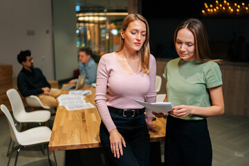 Portrait of two pretty young businesswomen working, talking together, reading paper document, having informal meeting on background of startup business team chatting at desk on background.