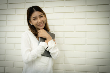 Beautiful asian female teacher feeling happy after finishing class teaching she hugged the tablet and looking to the camera with a smiling face against a white brick wall background.