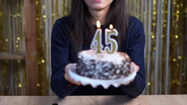 Birthday celebration concept. Happy woman holding birthday cake with burning candles number 45 and looking at the camera at home. Point of view or POV.