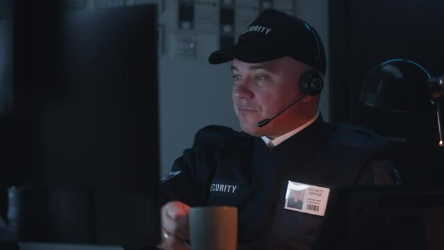 Sleepy security officer in headphones sits on workplace and looks view from security cameras displayed on computer monitor. Man drinks cup of coffee. Monitoring and tracking systems. CCTV technology.