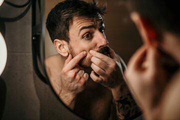 Shot of a young man examining his skin in the mirror at home. Health, skincare and man popping acne in face