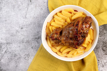 Roasted pork knuckle with garlic and potatoes in a white baking dish on a dark grey background. Top view, flat lay
