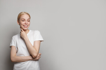 Attractive blonde female model in white t-shirt looking aside and smiling against grey studio wall banner background