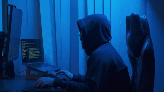 Hacker in hood cracking code using laptop from his dark hacker room