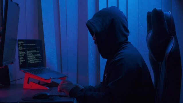 Hacker in hood cracking code using laptop from his dark hacker room