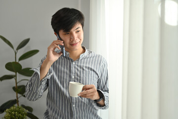 Portrait of smiling asian businessman holding coffee cup and talking on mobile phone at bright home office