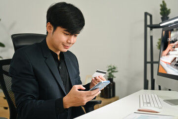 Smiling millennial asian man holding credit cad and using mobile phone smartphone for online banking transaction