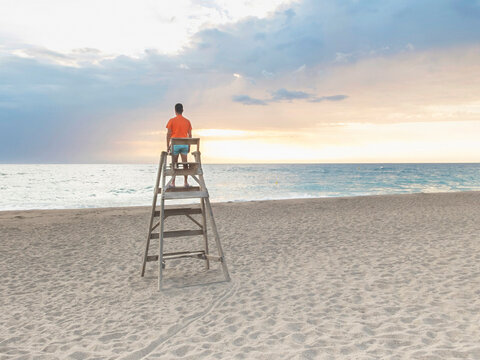 Rear view of man standing on lifeguard hut at beach against cloudy sky during sunset