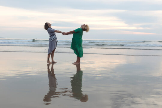 Full length of happy female friends holding hands while standing on shore at beach during sunset