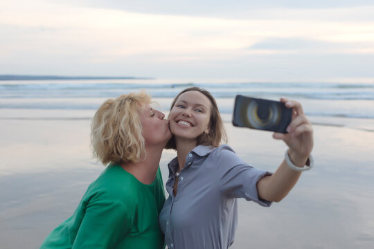 Cheerful woman taking selfie with smart phone while being kissed by friend at beach during sunset
