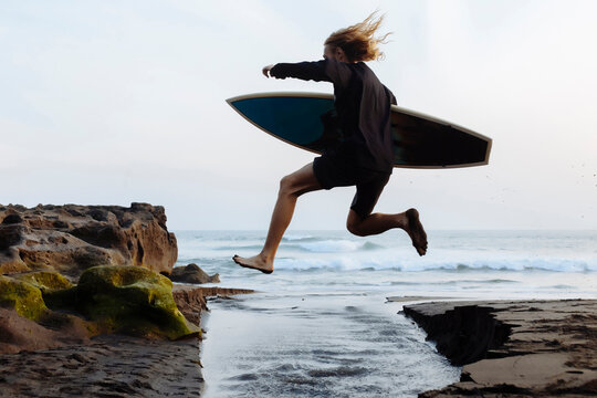 Full length of excited man carrying surfboard while jumping over water at beach