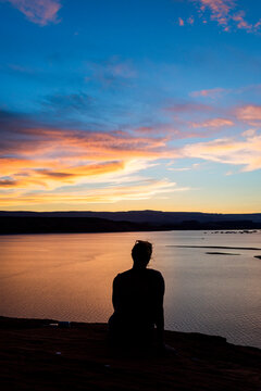 Silhouette woman sitting by river against dramatic sky during sunset
