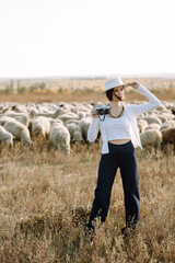 Young woman with a camera in front of a herd of sheep. Nature, fashion, vacation and lifestyle.