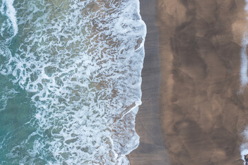 View of a beach from a drone. Turquoise water with waves on one side and snow-covered sand on the other.