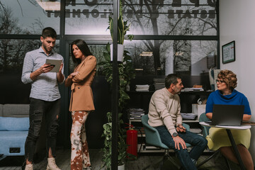 Young business couple standing and looking at the tablet. Discussing business. Their middle-aged couple colleagues are sitting and having serious conversation