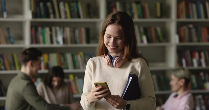 Cheerful pretty young high school student girl using app on smartphone in college library, typing, reading good news, turning to window, smiling, getting happy, thinking over message