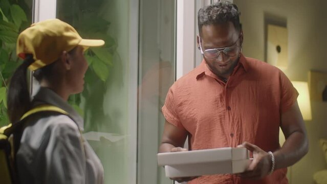Cheerful African American man opening door and receiving pizza boxes from female courier while using food delivery service