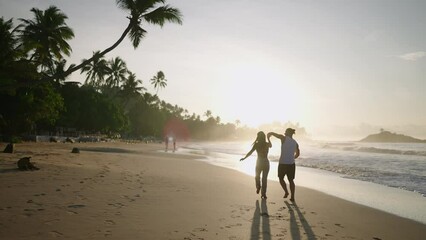 Young happy couple holding hands and running and fooling around on the beach together enjoying summer back view. Boyfriend and girlfriend having fun at the seaside chasing each other at sunrise.
